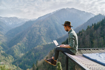 man sitting on a hammock suspended over a valley working on laptop