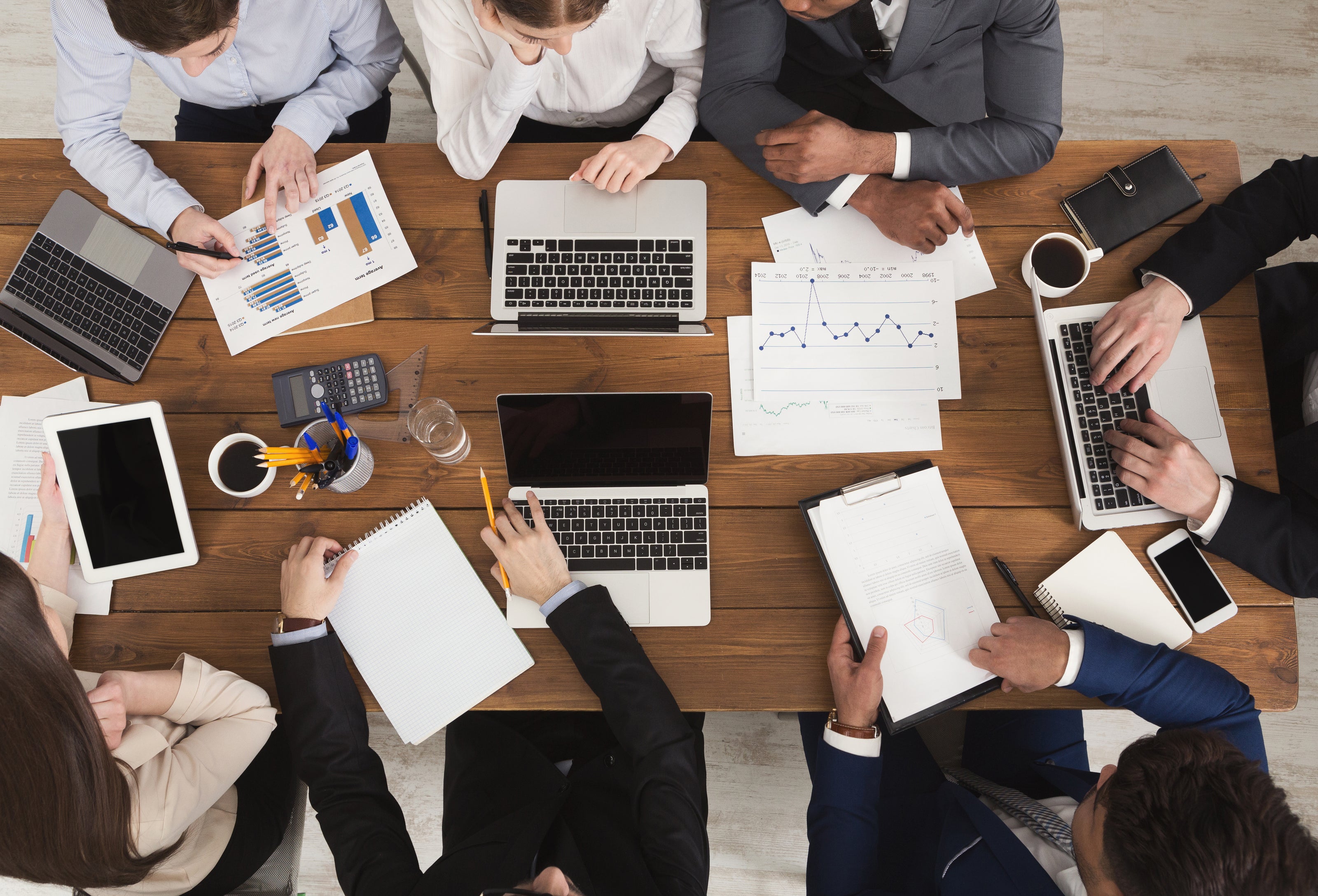Group of people working together at a table with laptops, tablets, and papers.